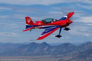 The red white and blue MSU Denver game bird aircraft soars in the air on a sunny day with the rocky mountains in the background.